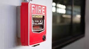Red fire alarm pull station mounted on a wall, with a window in the background reflecting light.